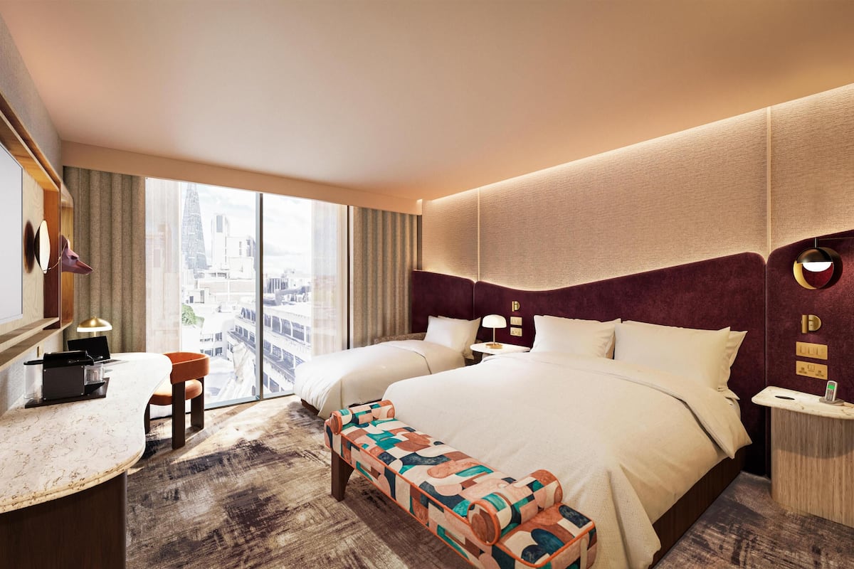 Habitación de hotel con una cama grande, un escritorio, un banco y vistas de la ciudad, con un paisaje urbano a través de la ventana.