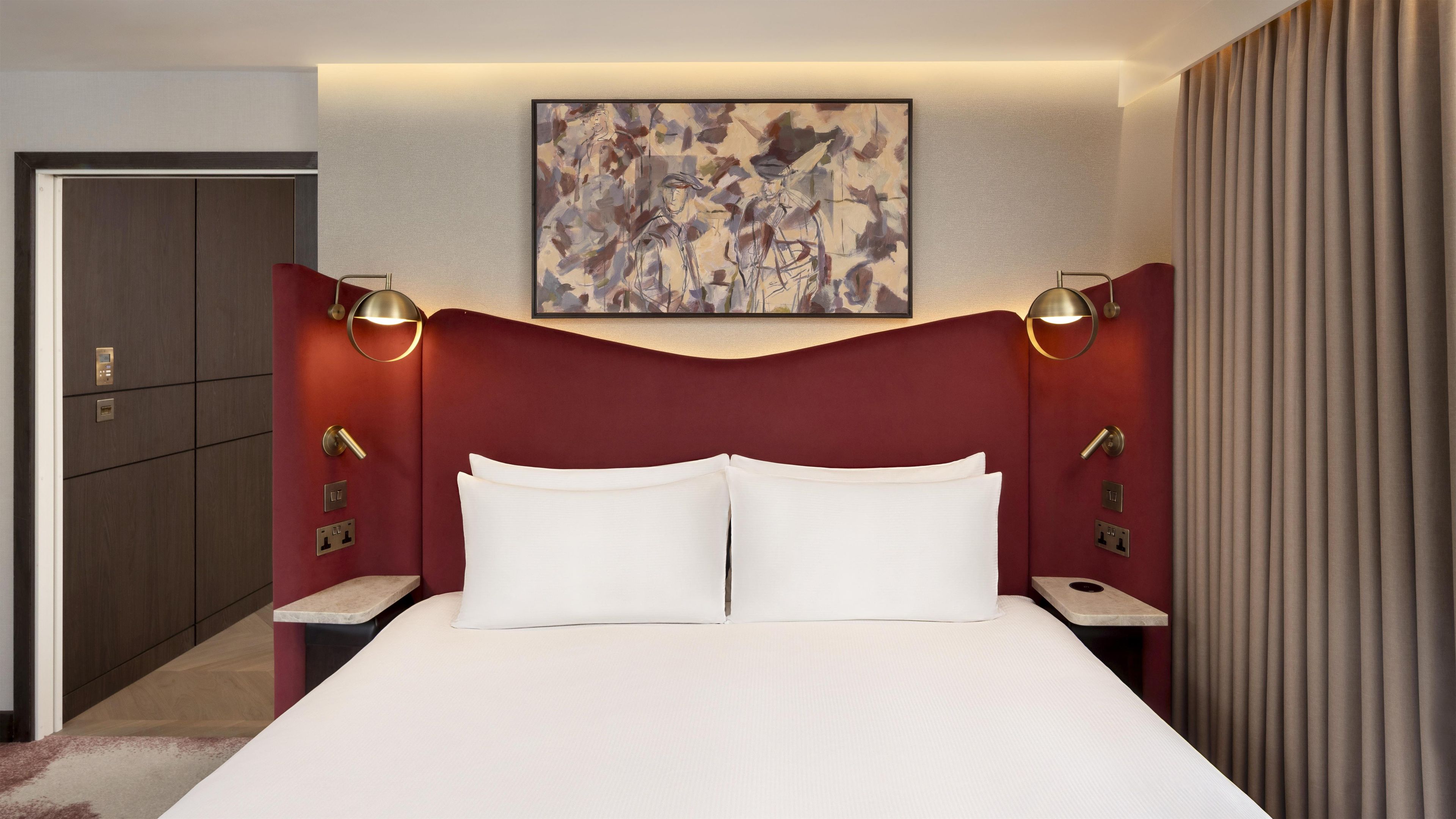 A hotel room with a large bed, two bedside tables, a headboard, a painting on the wall, and a door to the left.