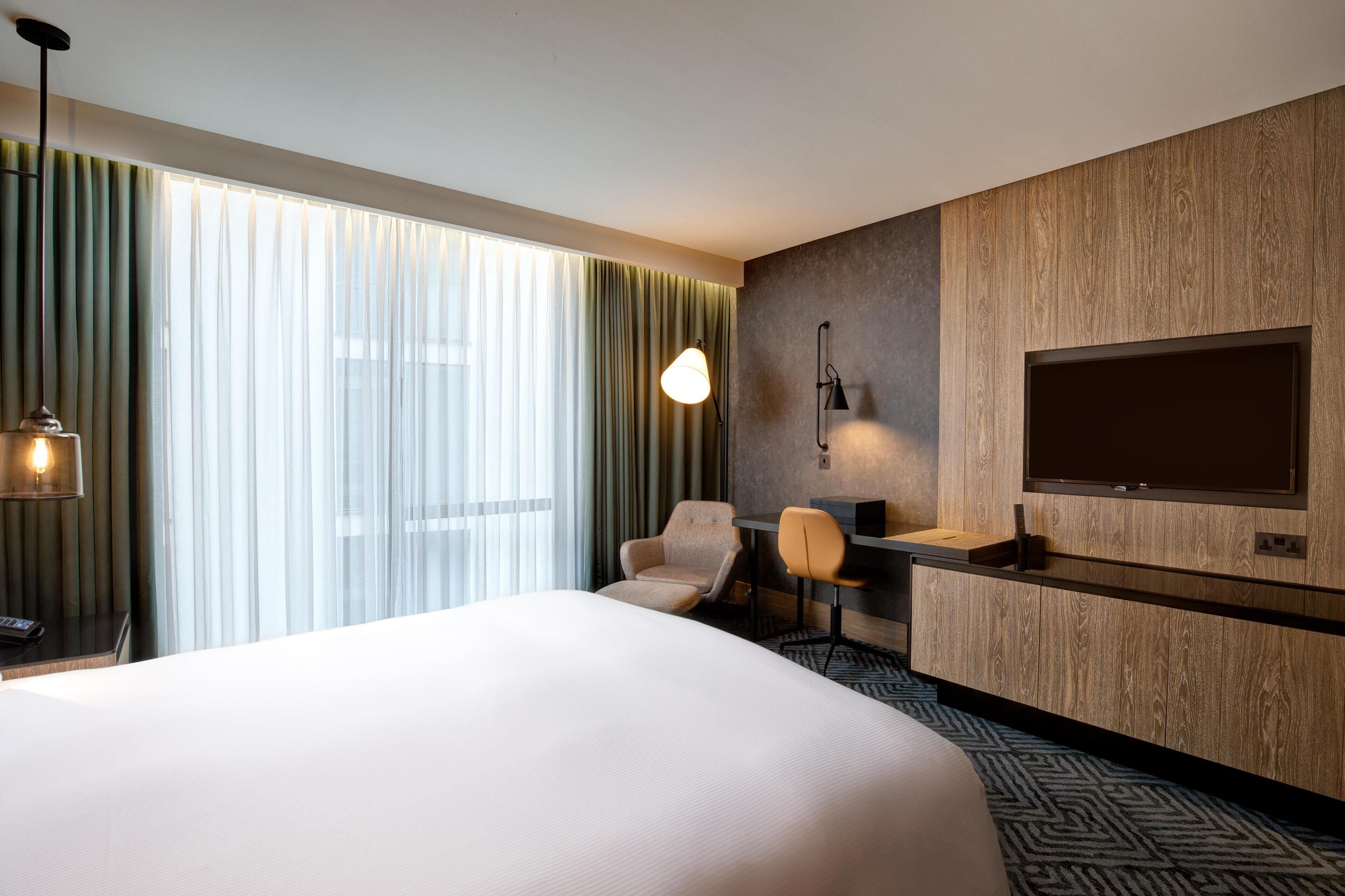 Ein Hotelzimmer mit einem großen Bett, einem Schreibtisch mit Stuhl, einem an der Wand befestigten Fernseher und einem großen Fenster mit Vorhängen.