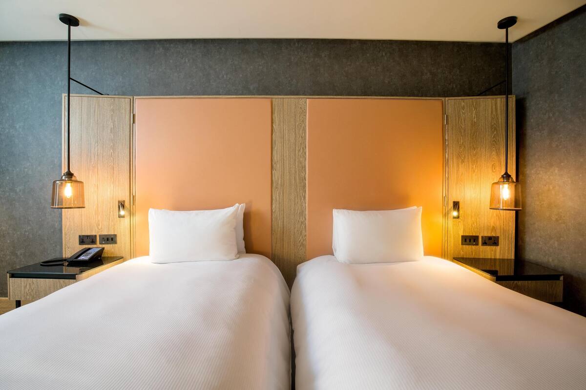 Habitación de hotel con dos camas, cabecero de madera, lámparas de noche, un teléfono en la mesita y una lámpara de pared.