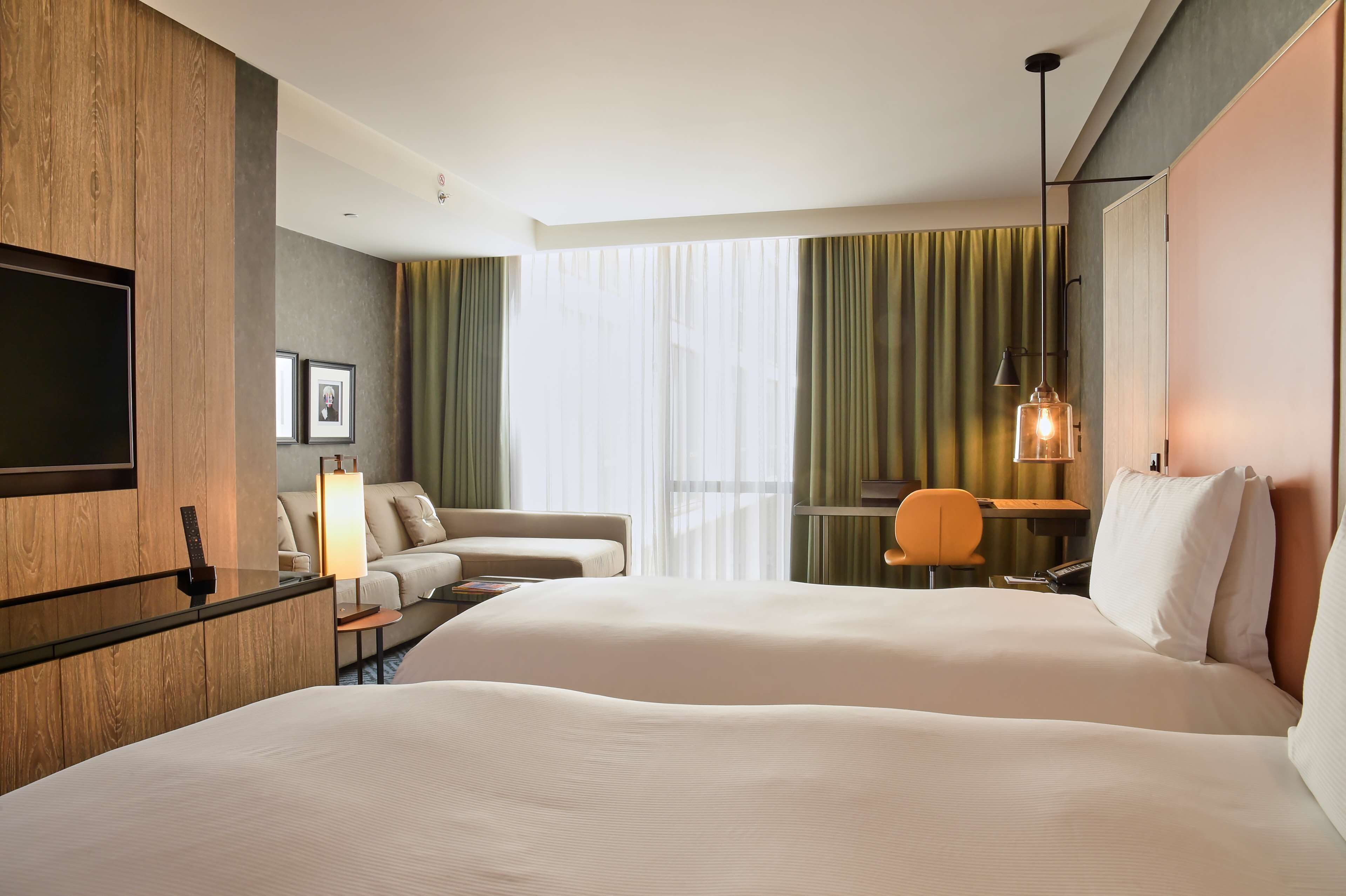 A hotel room with two beds, a desk, a chair, a TV, and a large window with curtains.