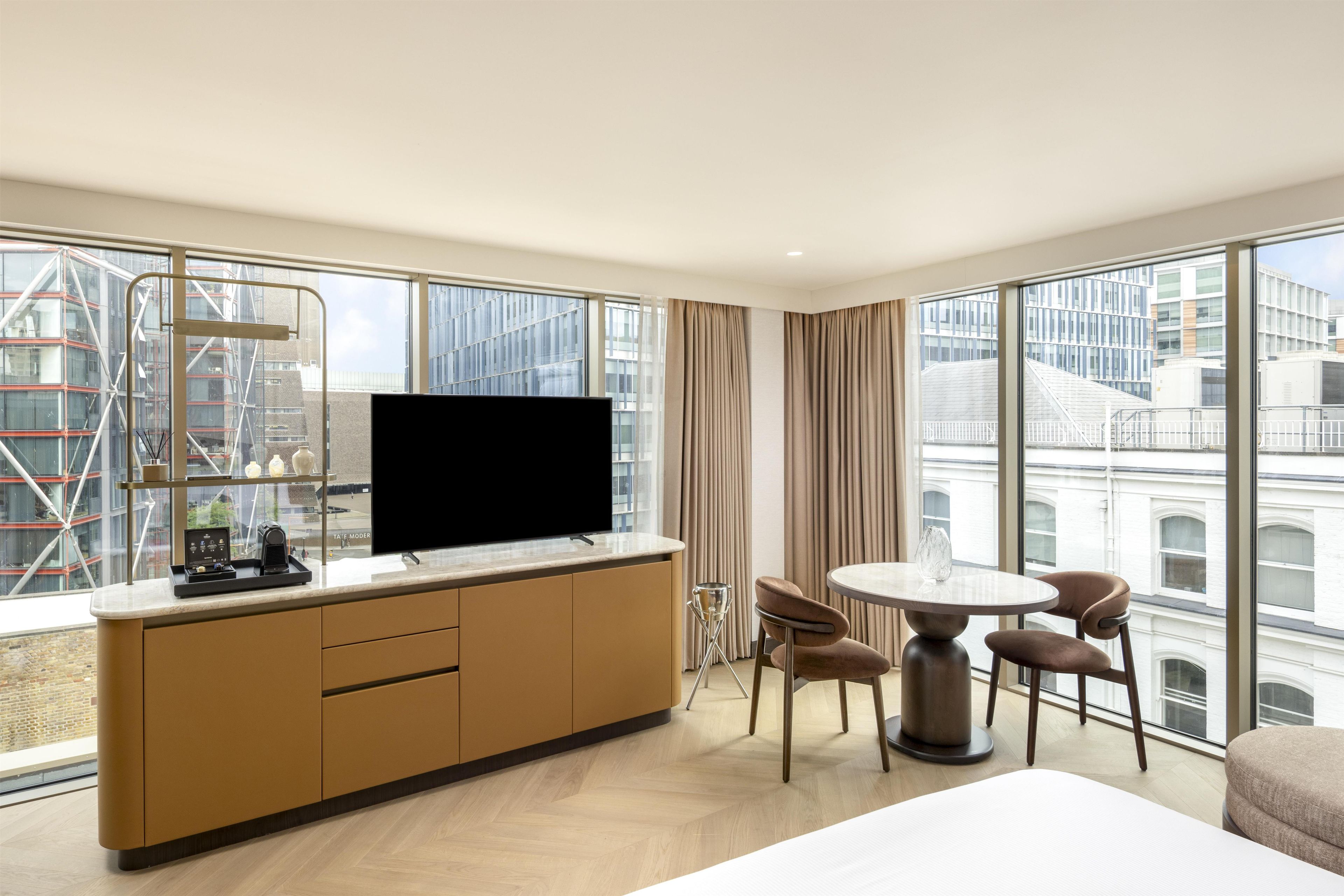 Ein Hotelzimmer mit großem Fenster, einem Flachbildfernseher, einem Esstisch mit Stühlen und einem Bett.