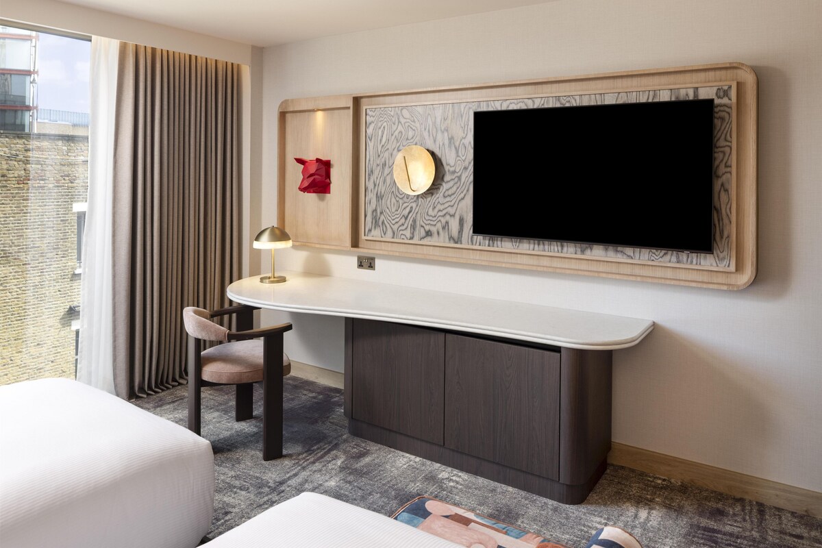 Habitación de hotel con escritorio, una silla, televisor montado en la pared y ventana con cortinas.