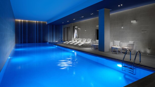 A modern indoor pool with a blue-lit ceiling, lounge chairs, and a seating area with tables. - Hilton London Bankside (London)