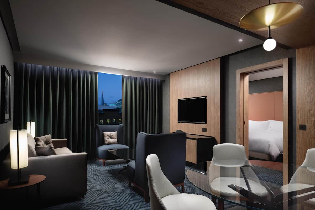 Una habitación de hotel moderna con sofá, sillones, una mesa de vidrio, televisión y cama.