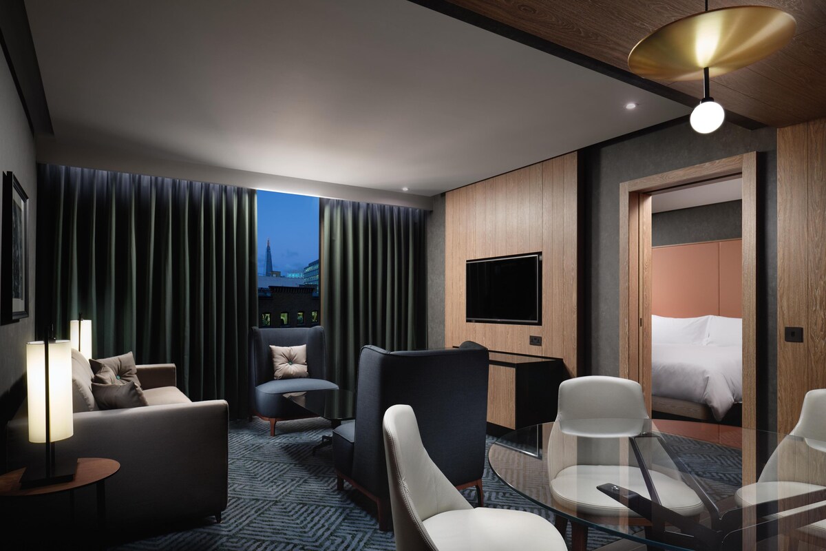 Una habitación de hotel moderna con sofá, sillones, una mesa de vidrio, televisión y cama.