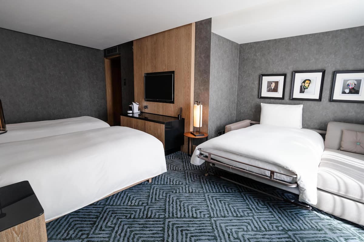 Habitación de hotel con dos camas, un televisor, un escritorio con lámpara y cuadros en la pared.
