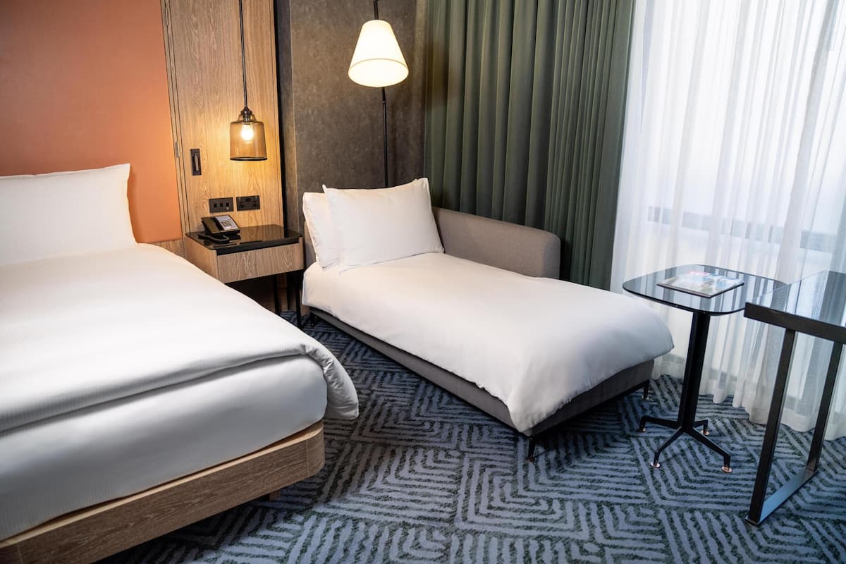 Habitación de hotel con dos camas, una mesita de noche, una lámpara, una pequeña mesa y vistas al exterior a través de cortinas translúcidas.