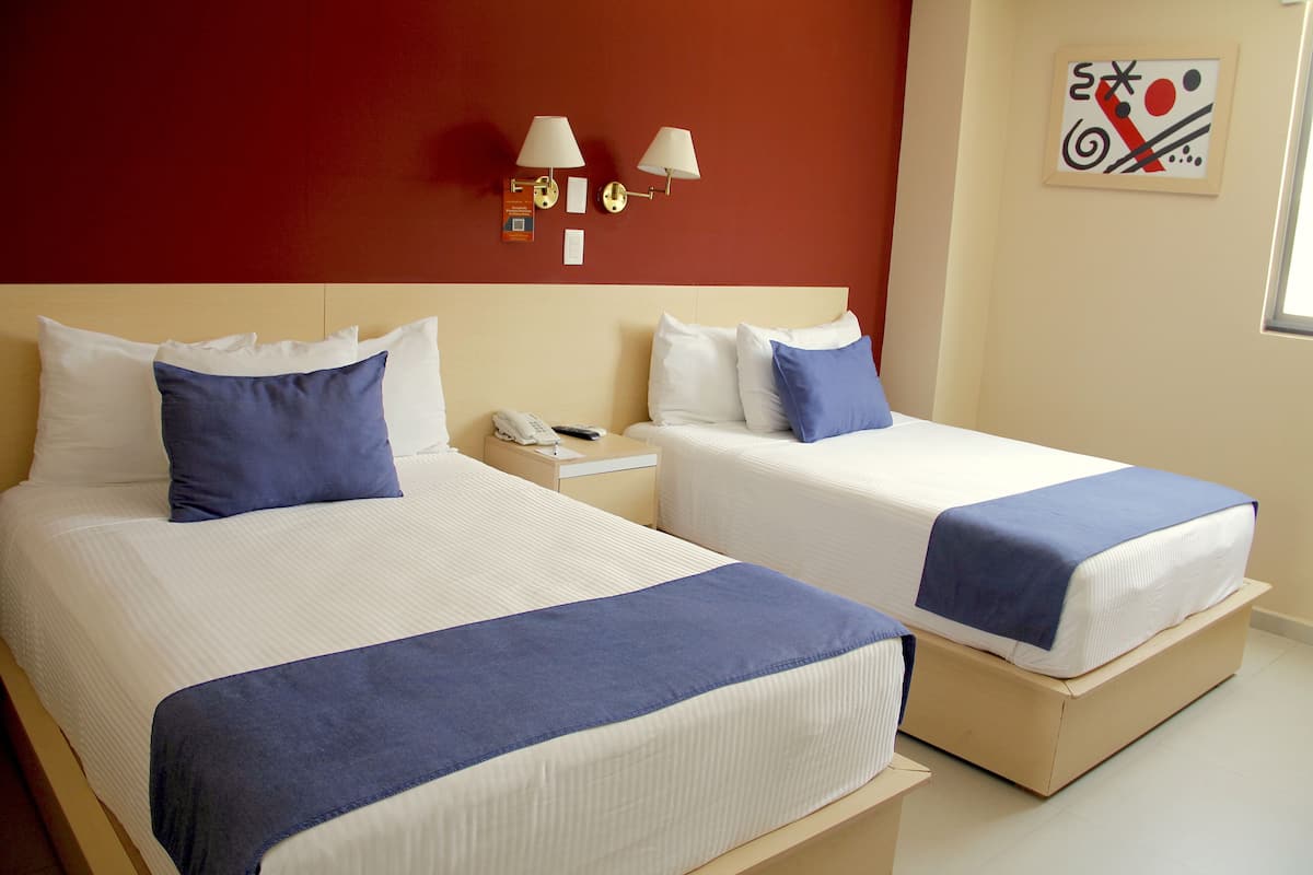 Two beds with white linens and blue accents, a nightstand with a phone, and wall-mounted lamps.