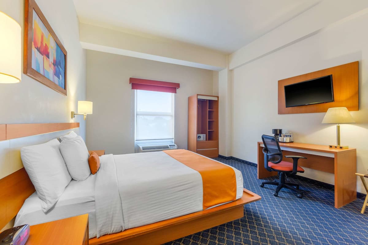 A hotel room with a bed, desk, chair, TV, and a window with a red curtain.