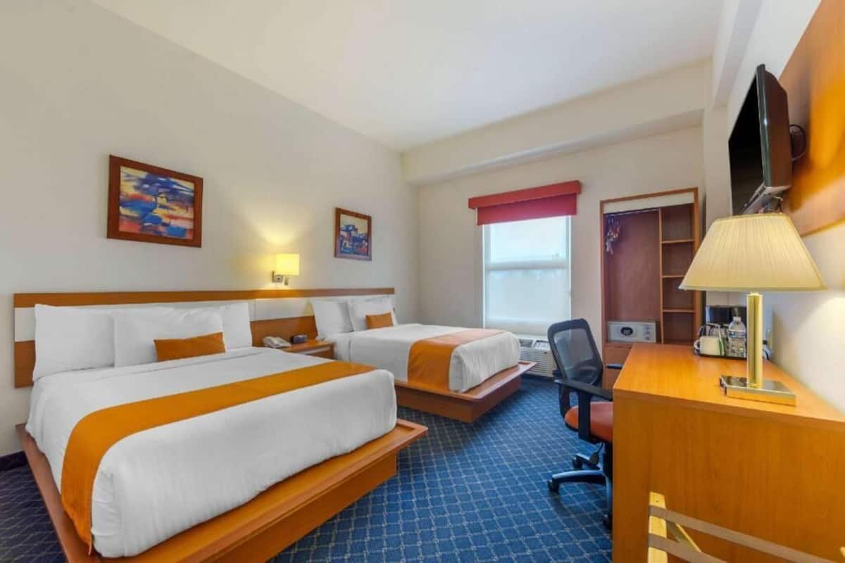 A hotel room with two beds, a desk, a chair, a TV, and a window with a red curtain.