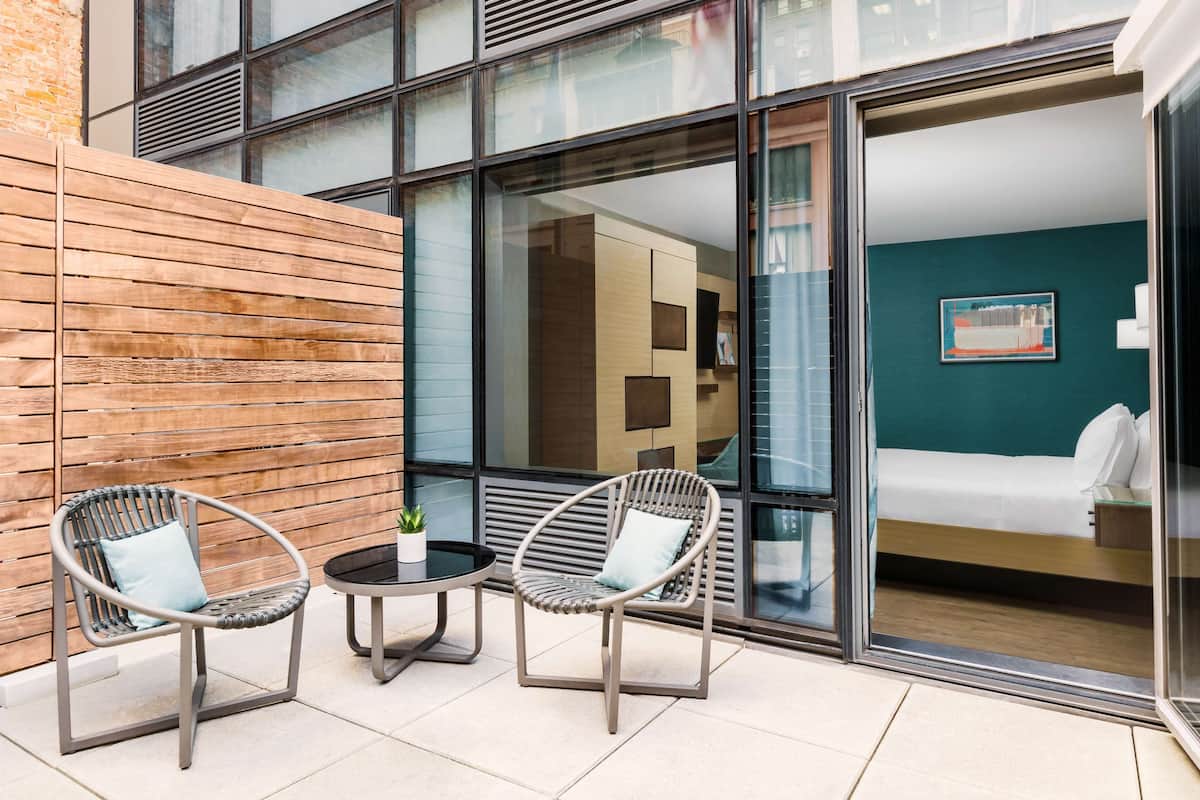 Une chambre d’hôtel moderne dotée d’une grande porte vitrée, de deux fauteuils en osier, d’une petite table et offrant une vue sur le paysage urbain.