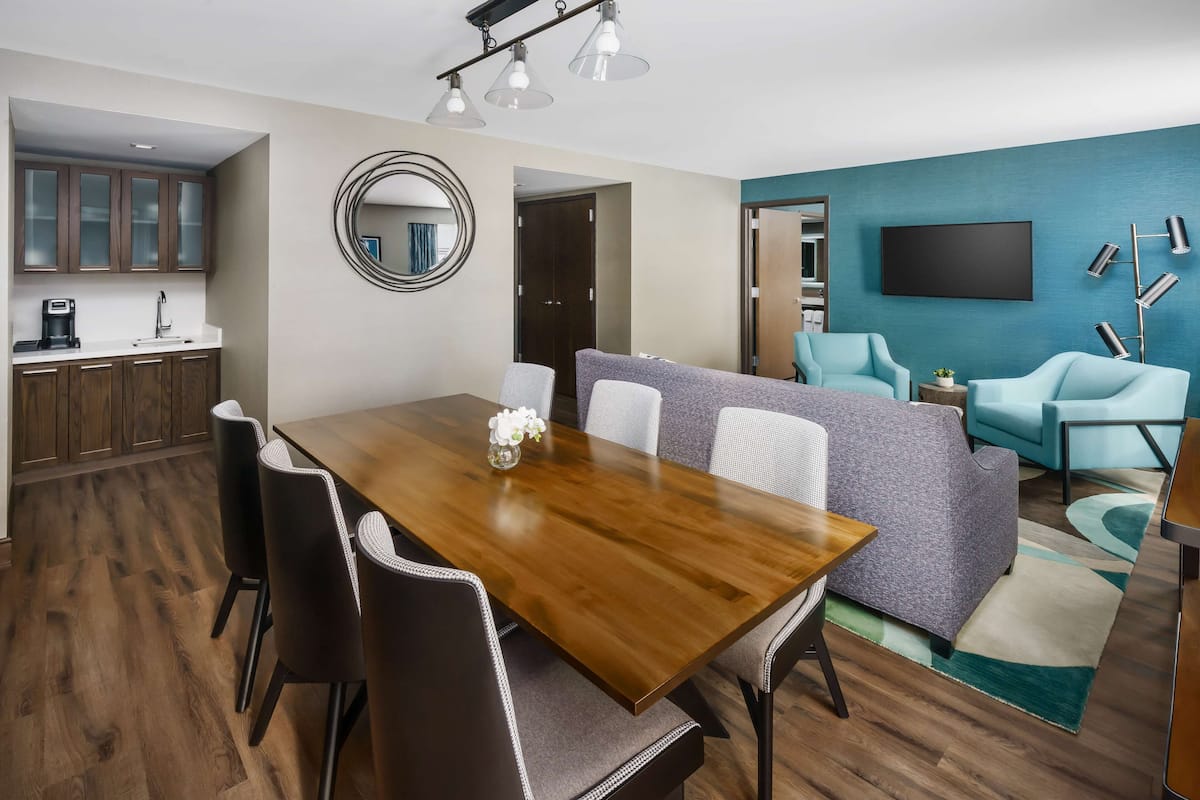 Un salon moderne avec une table à manger en bois, un canapé gris et des fauteuils turquoise. Un téléviseur à écran plat et une lampe sur pied sont fixés au mur.
