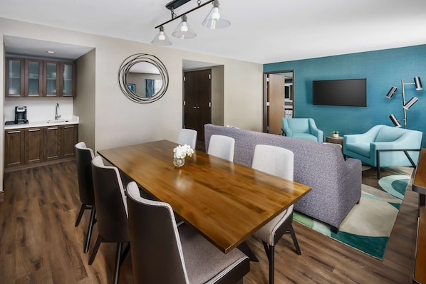 A modern living room with a wooden dining table, grey sofa, and teal armchairs. A flat-screen TV and a floor lamp are mounted on the wall.