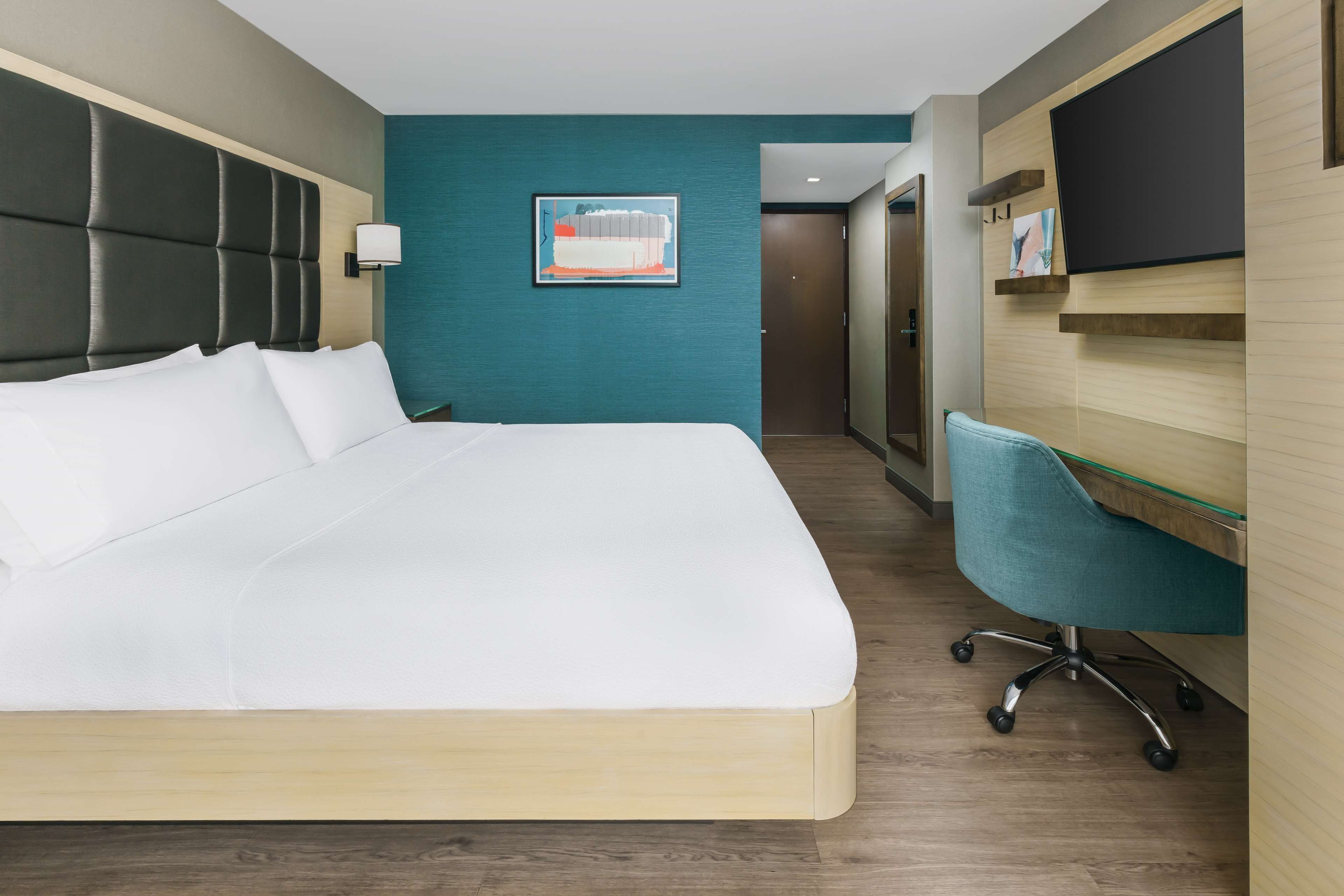 A hotel room with a large bed, a desk with a chair, a TV mounted on the wall, and a door leading to another room.