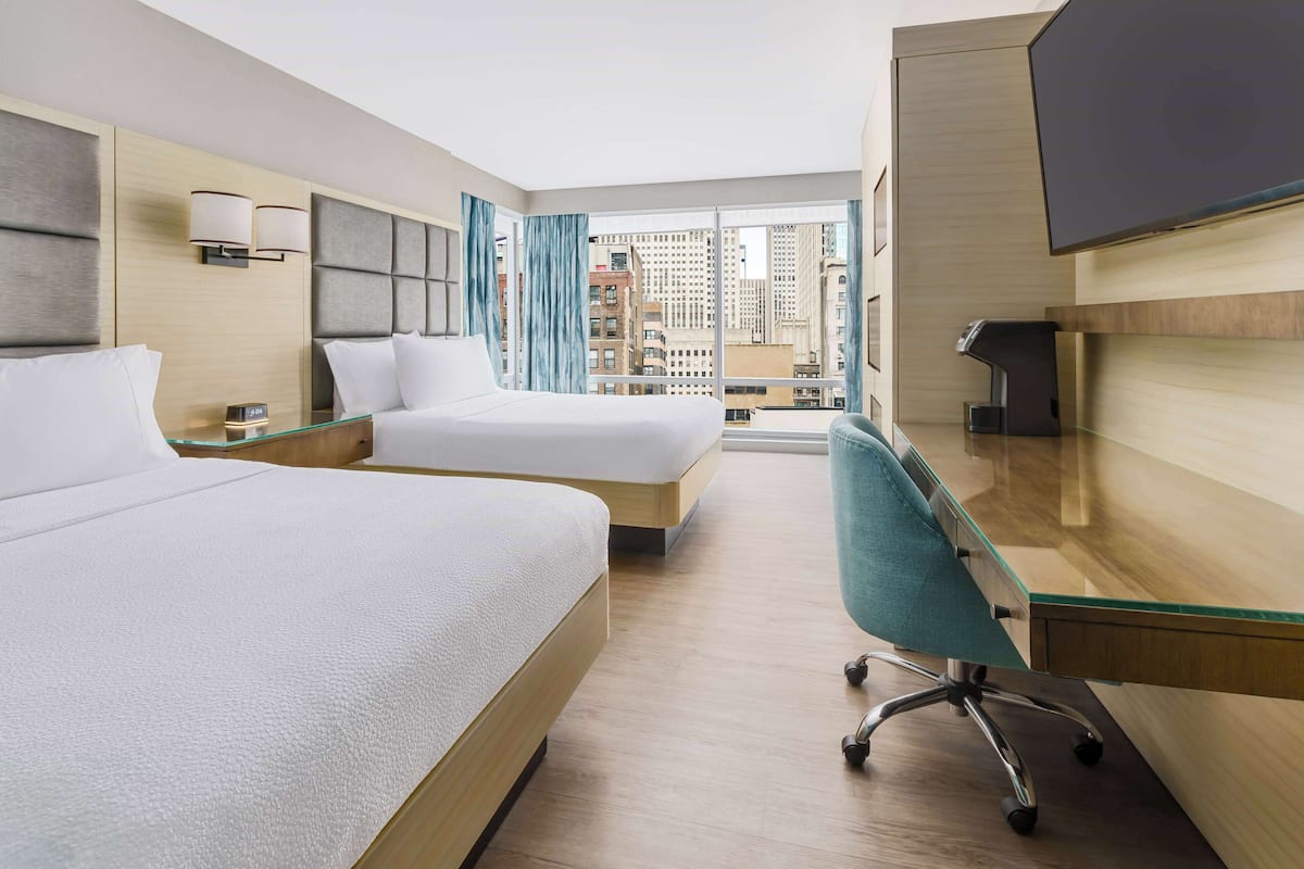 Une chambre d’hôtel avec deux lits, un bureau, une chaise, une télévision et une grande fenêtre donnant sur la ville.