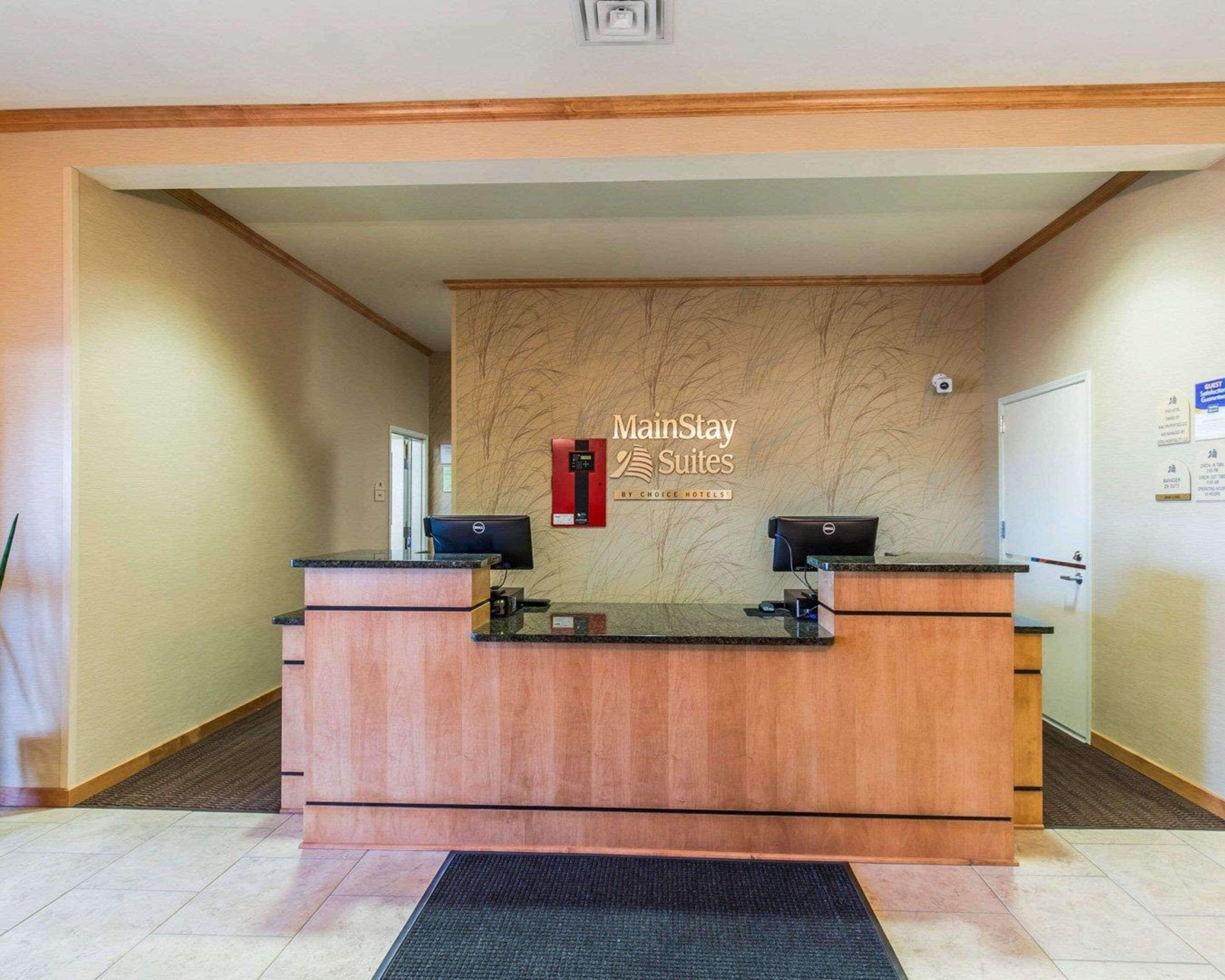 A reception desk with a wooden finish, two computer monitors, a fire alarm, and a sign for MainStay Suites by Choice Hotels.