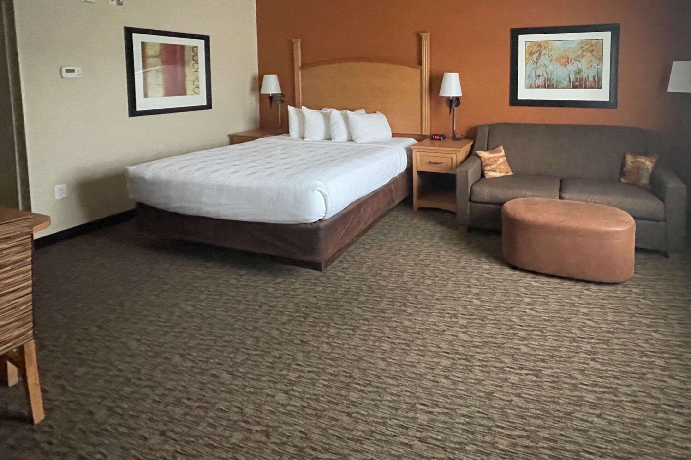 Quarto de hotel com uma cama grande, um sofá, um pequeno banquinho e duas mesas de cabeceira com abajures.