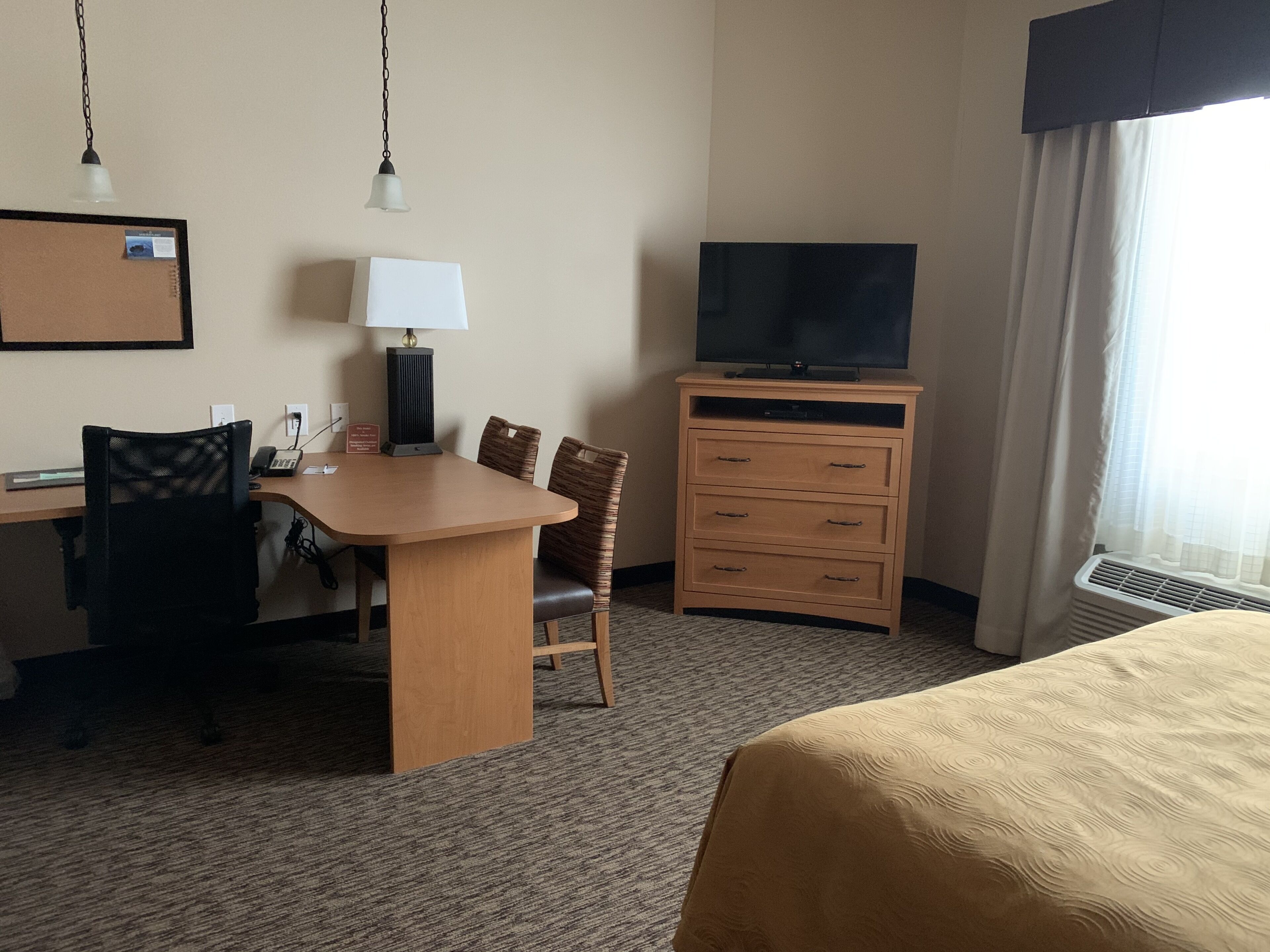 Habitación de hotel con cama, escritorio con silla, televisor sobre un soporte, lámpara y cómoda.