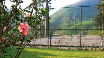 A tennis court with a net, surrounded by a fence and a grassy area with a flower bush in the foreground.