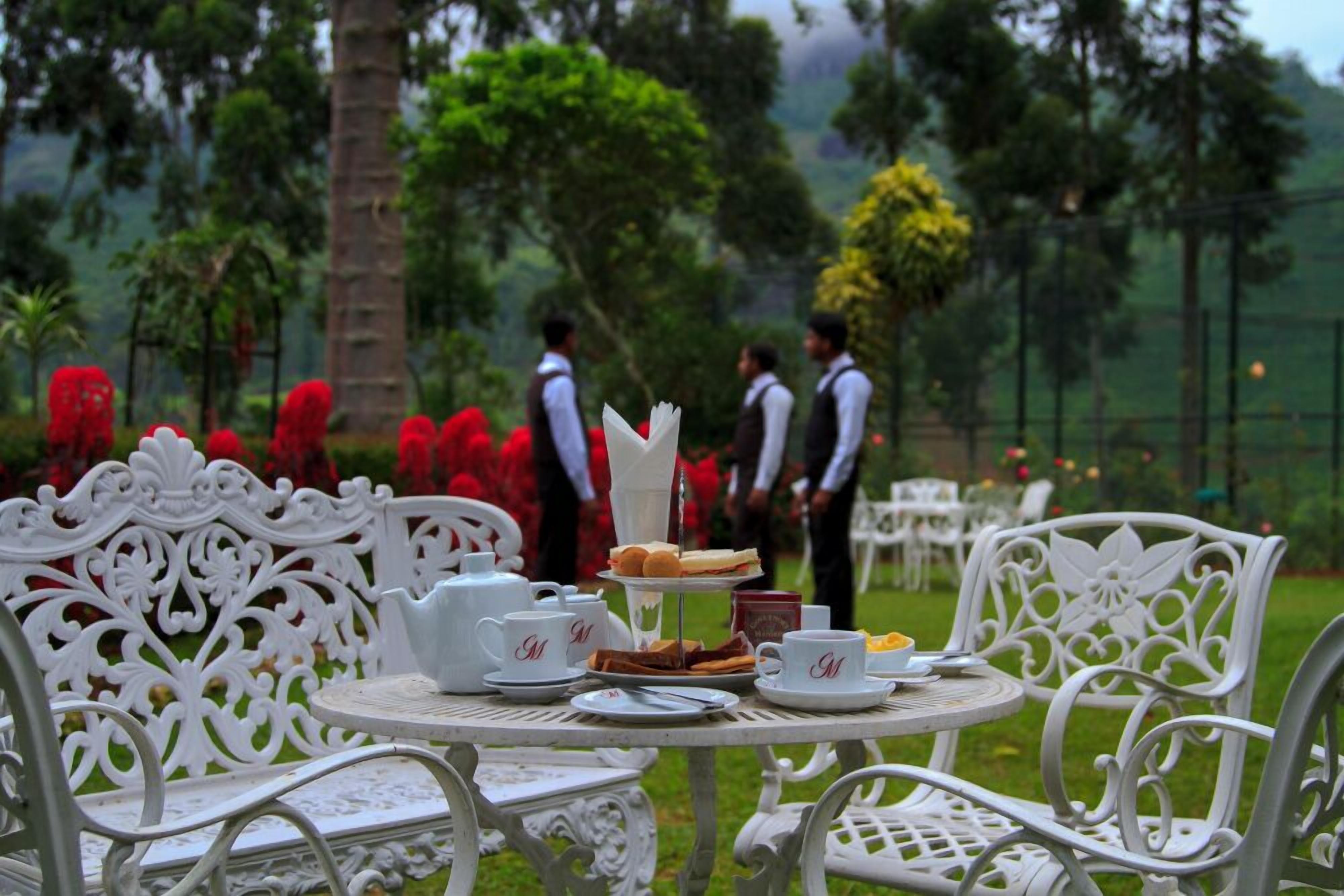 An outdoor dining area with white ornate chairs and tables, a tiered dessert stand, and a view of a lush garden with palm trees and red flowers.