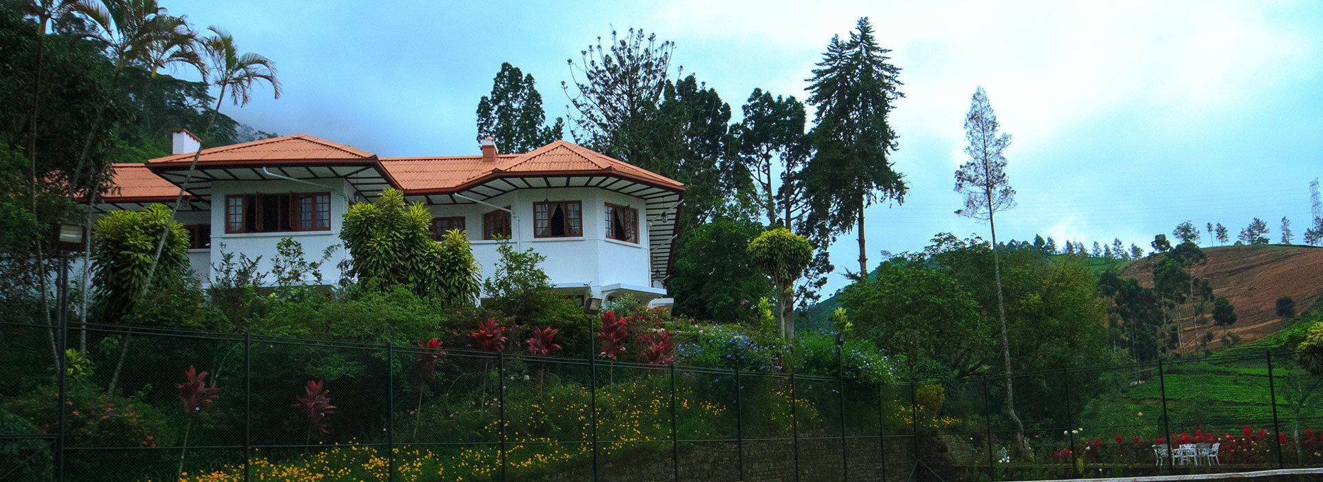 Una casa de dos pisos con techo rojo, rodeada de abundante vegetación y una cerca.