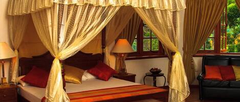 A four-poster canopy bed with two bedside lamps, a sofa, and a view of greenery outside.