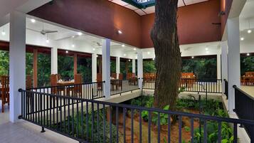 A covered outdoor dining area with tables and chairs, surrounded by greenery and a central tree.