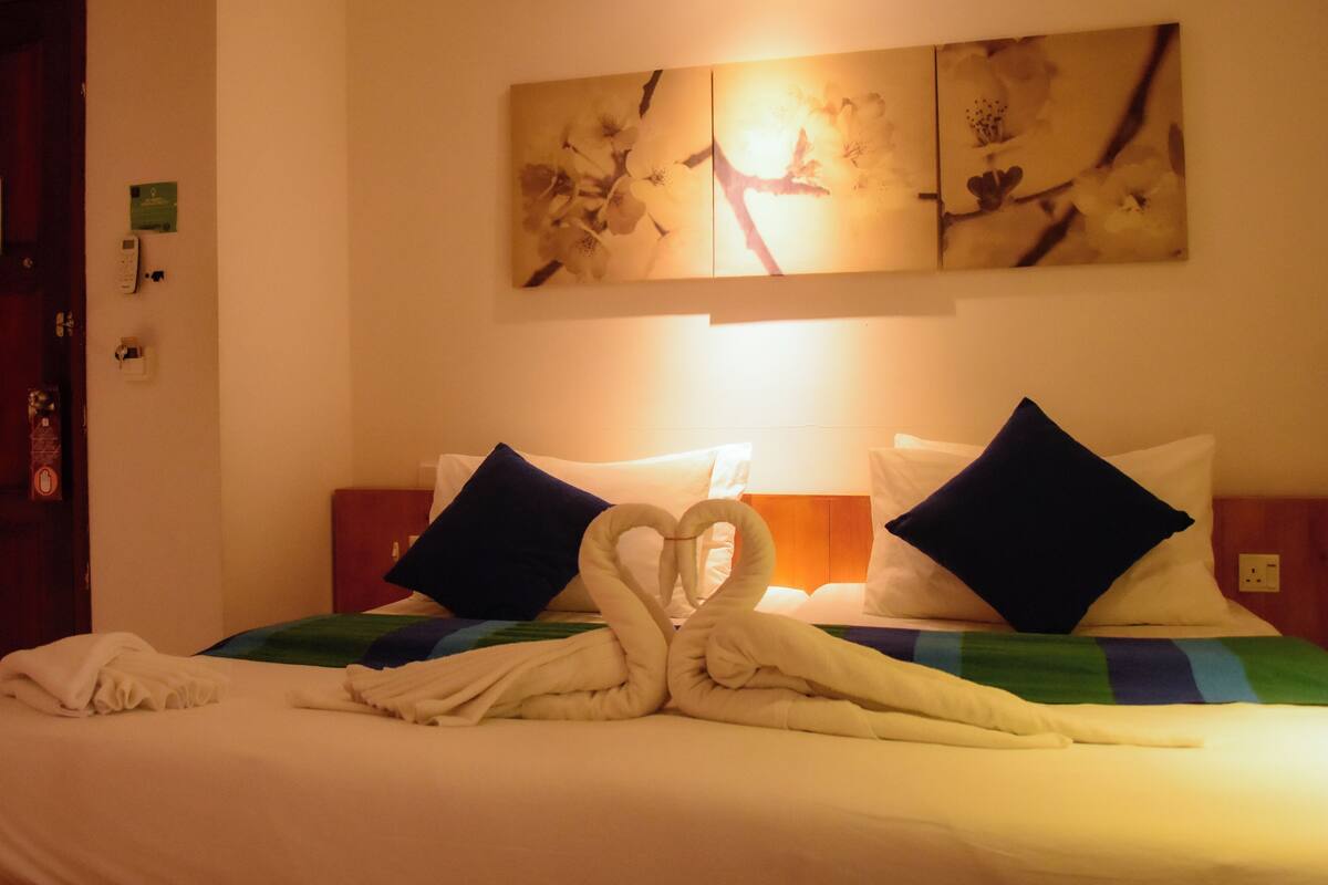 A hotel room with a bed, pillows, a painting, and a wall-mounted light.