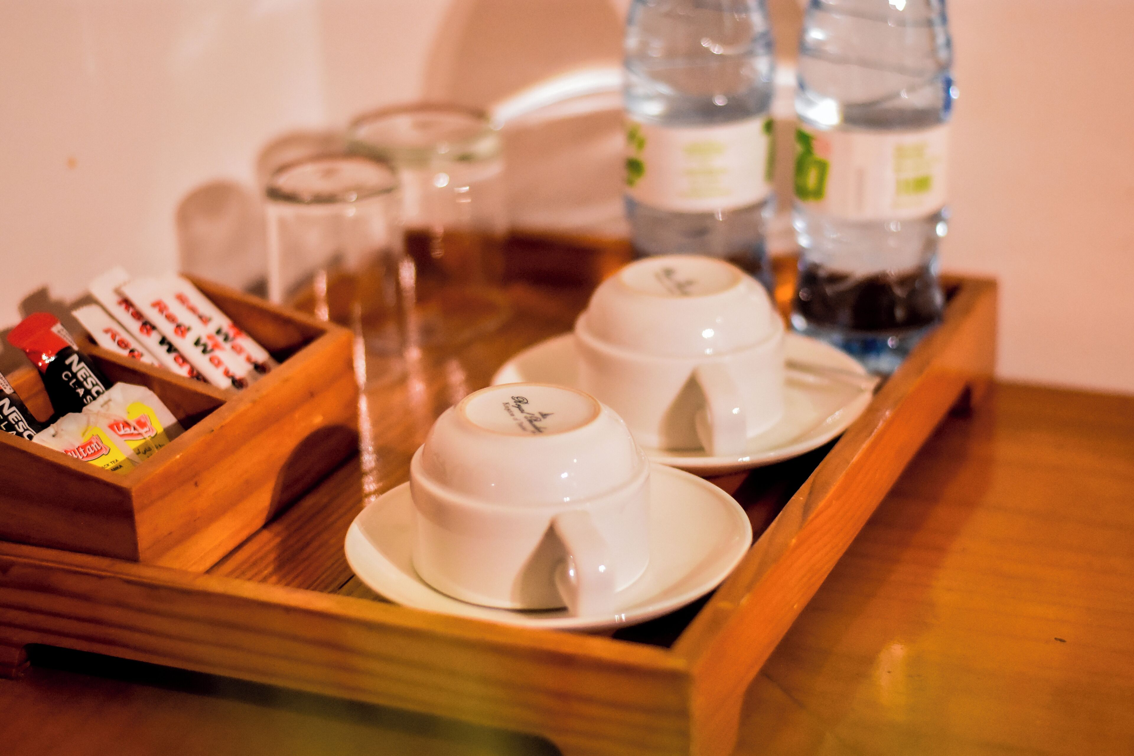 Una bandeja con dos tazas y platillos blancos, una caja de madera con varios objetos y botellas de agua.