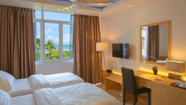A hotel room with two beds, a desk, a chair, a TV, and a large window with curtains. - Pine Lodge Maldives (Hulhumalé)