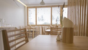 A dining area with wooden tables and chairs, large windows with blinds, and a white pendant light. - Pine Lodge Maldives (Hulhumalé)