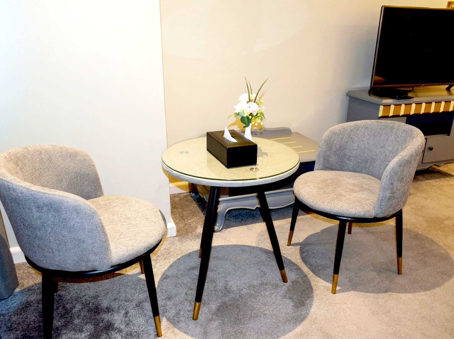 A room with two grey upholstered chairs, a small round table with a vase of flowers, a television on a stand, and a carpeted floor.