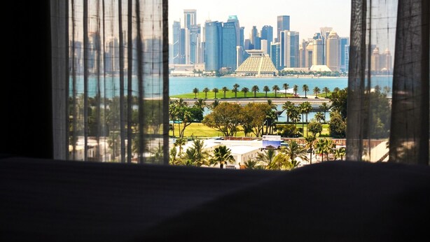 A view through a window with sheer curtains, featuring a city skyline, a body of water, and a park with palm trees. - Saraya Corniche Hotel (Doha)