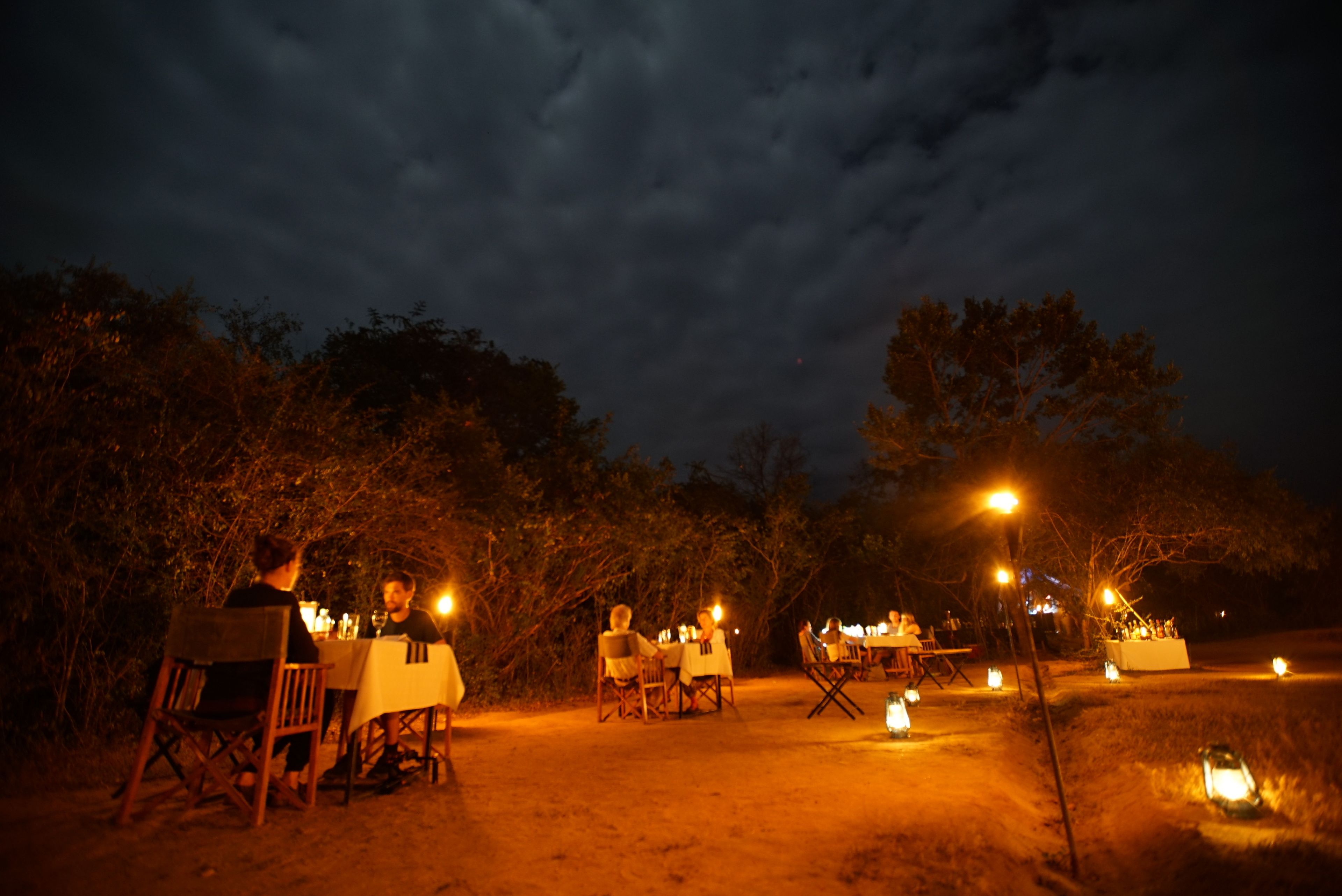 An outdoor dining area with tables and chairs set up under street lamps, surrounded by trees.