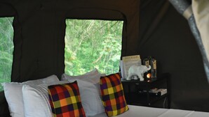 A tent with a bed, white pillows, colorful checkered pillows, a nightstand with a book and a candle, and a window with a forest view. - Big Game - Udawalawe by Eco Team (Thanamalvila)