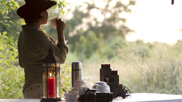 A person in a hat sitting at a table with a candle, a thermos, and various cups and mugs. - Big Game - Wilpattu by Eco Team (Pahala Maragahawewa)