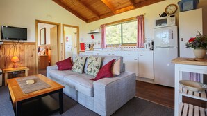 A cozy living room with a sofa, a coffee table, and a TV. There's a kitchen area with a fridge, microwave, and a window with curtains. - Buln Buln Cabins (Yanakie)