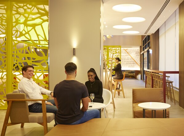 Un décor intérieur moderne où l’on voit un groupe de personnes assises autour d’une table, une femme debout près d’une échelle et un paravent décoratif jaune.