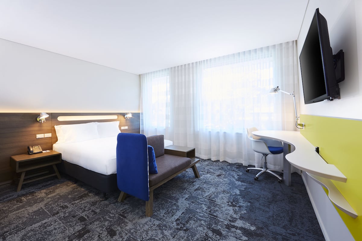 Habitación de hotel con cama, mesita de noche, lámpara, escritorio, silla y televisor montado en la pared.