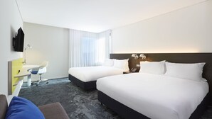 Quarto de hotel com duas camas, uma escrivaninha com cadeira, televisão de tela plana e uma janela grande com cortinas finas.