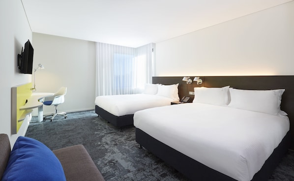 Een hotelkamer met twee bedden, een bureau met stoel, een flatscreen-tv en een groot raam met dunne gordijnen.
