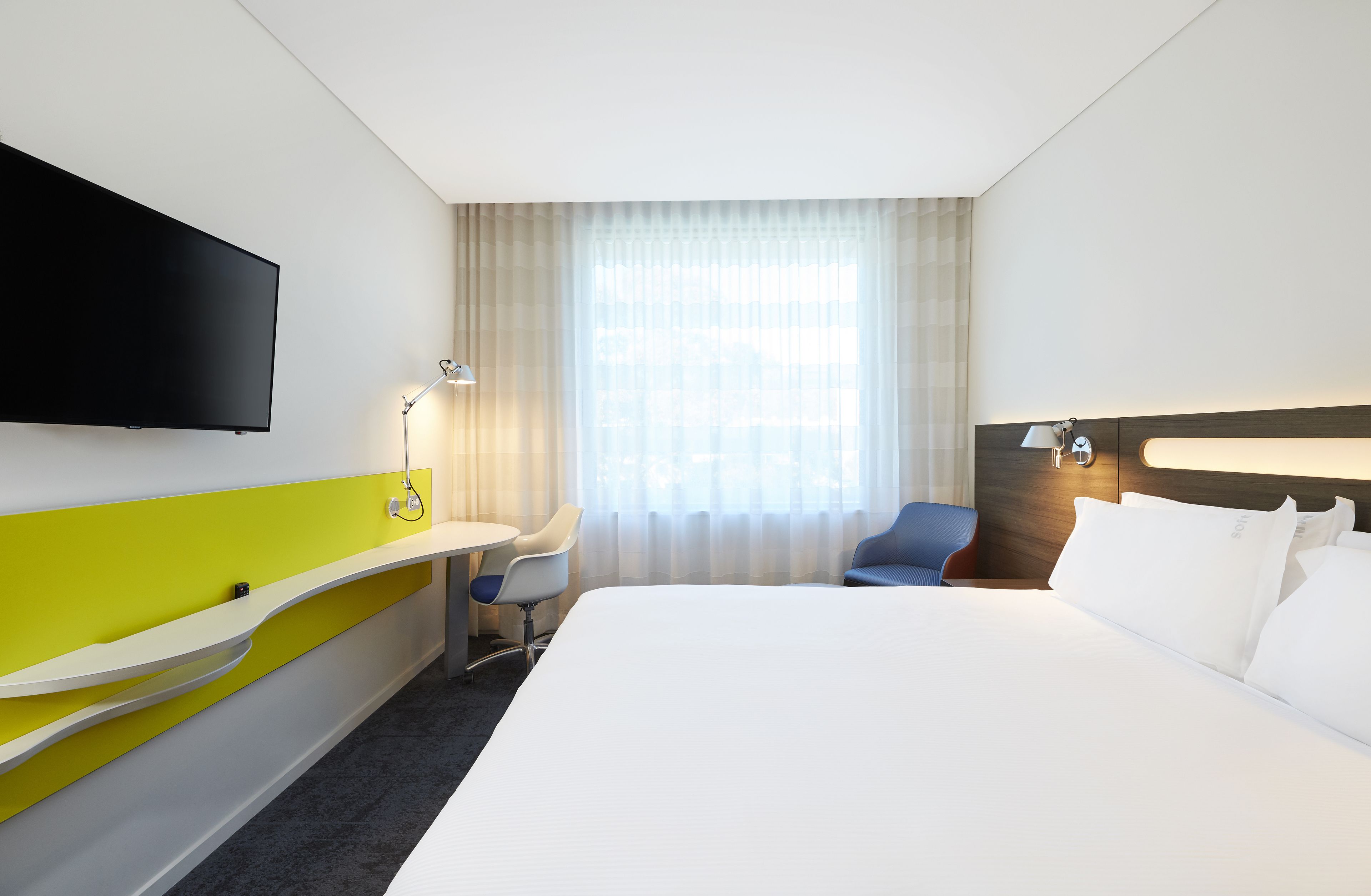 Habitación de hotel con una cama grande, un escritorio con una silla, un televisor de pantalla plana en la pared y un ventanal con cortinas translúcidas.