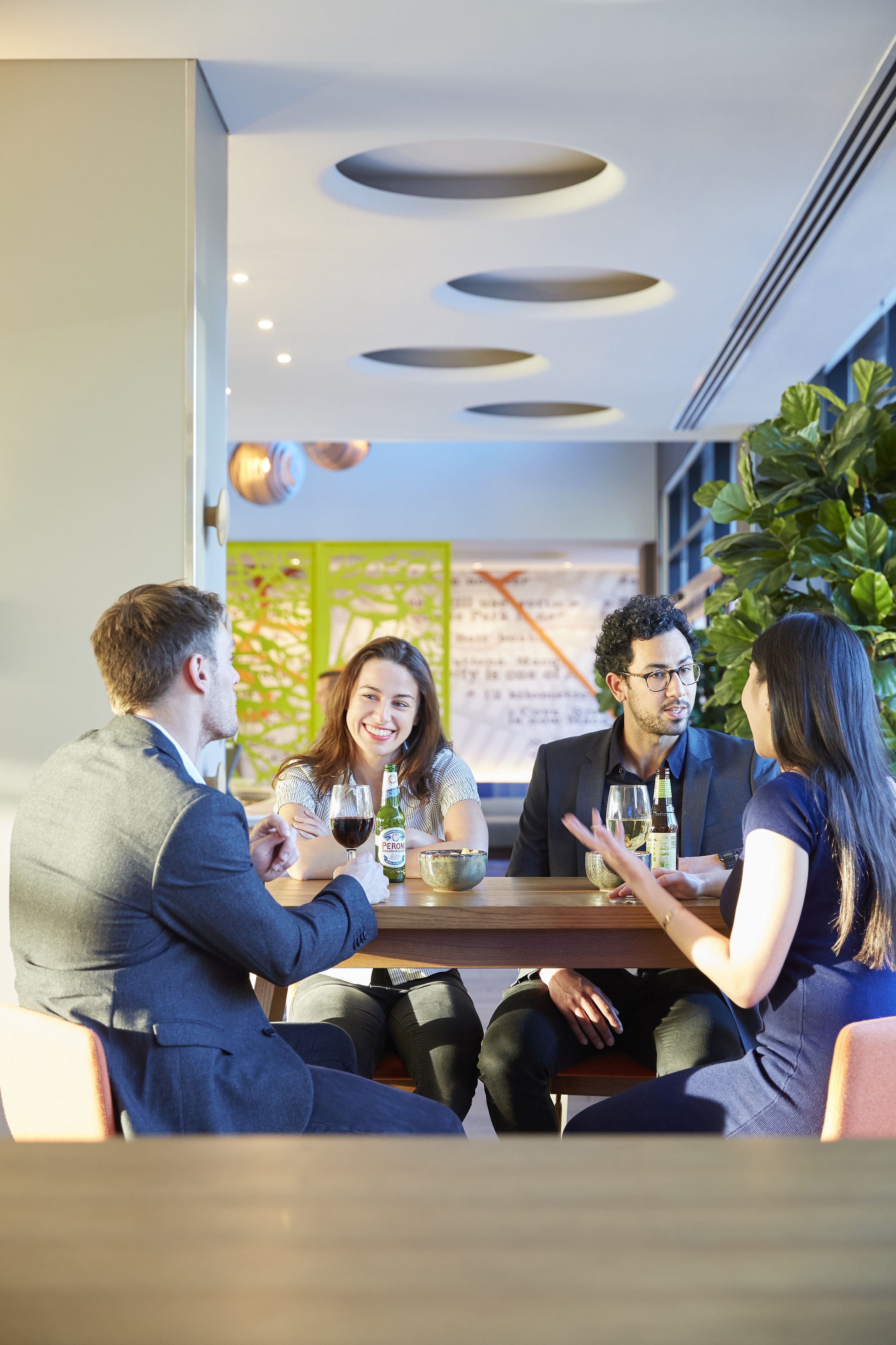 A group of four people seated around a table in a modern indoor setting with a decorative ceiling and a green plant in the background.