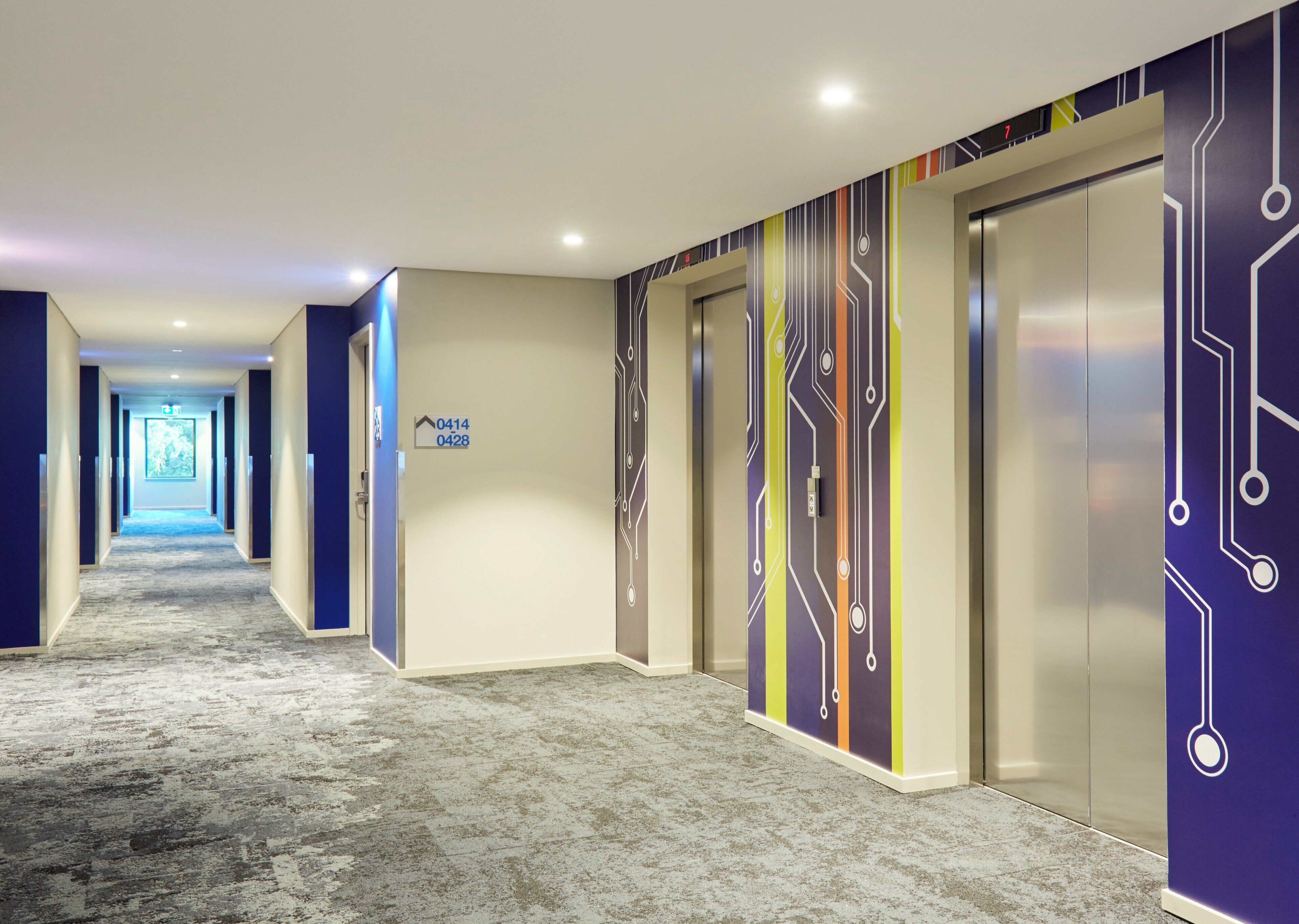 Un pasillo de hotel con piso alfombrado, un cartel con números de habitación y una pared con un diseño que recuerda a un circuito.