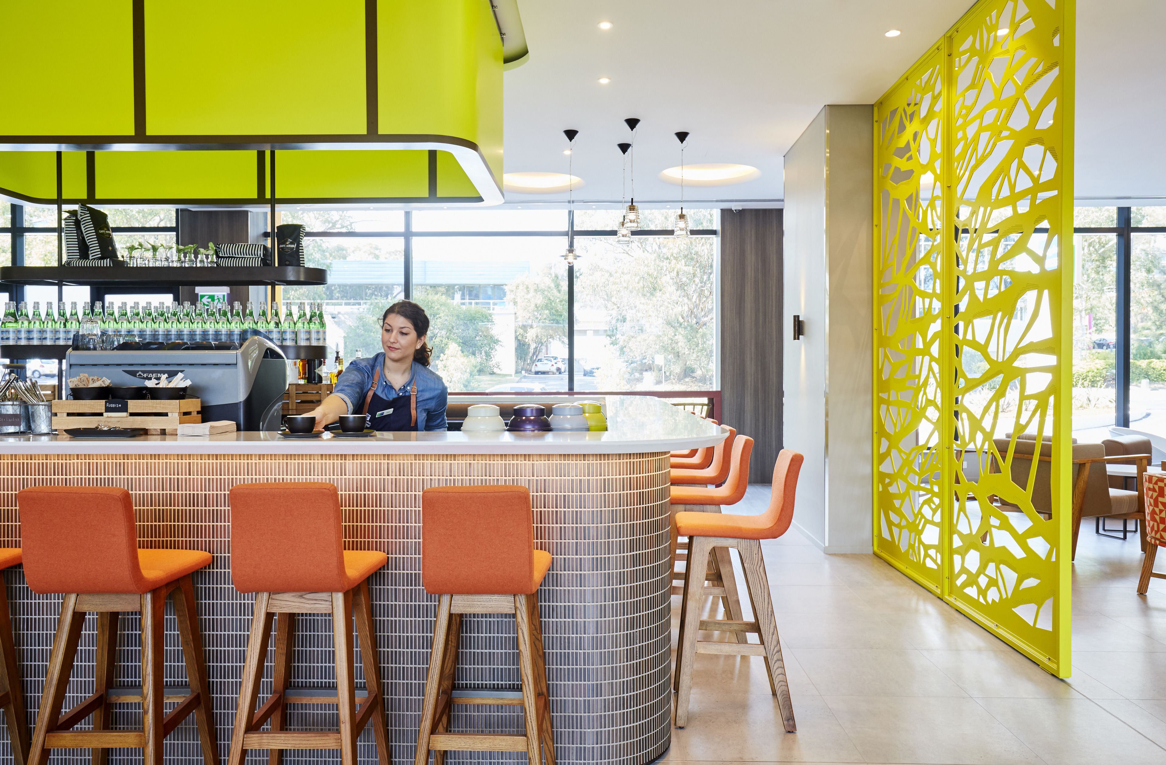 Un área de bar moderna con un barista preparando bebidas, taburetes de bar y un separador decorativo amarillo brillante.