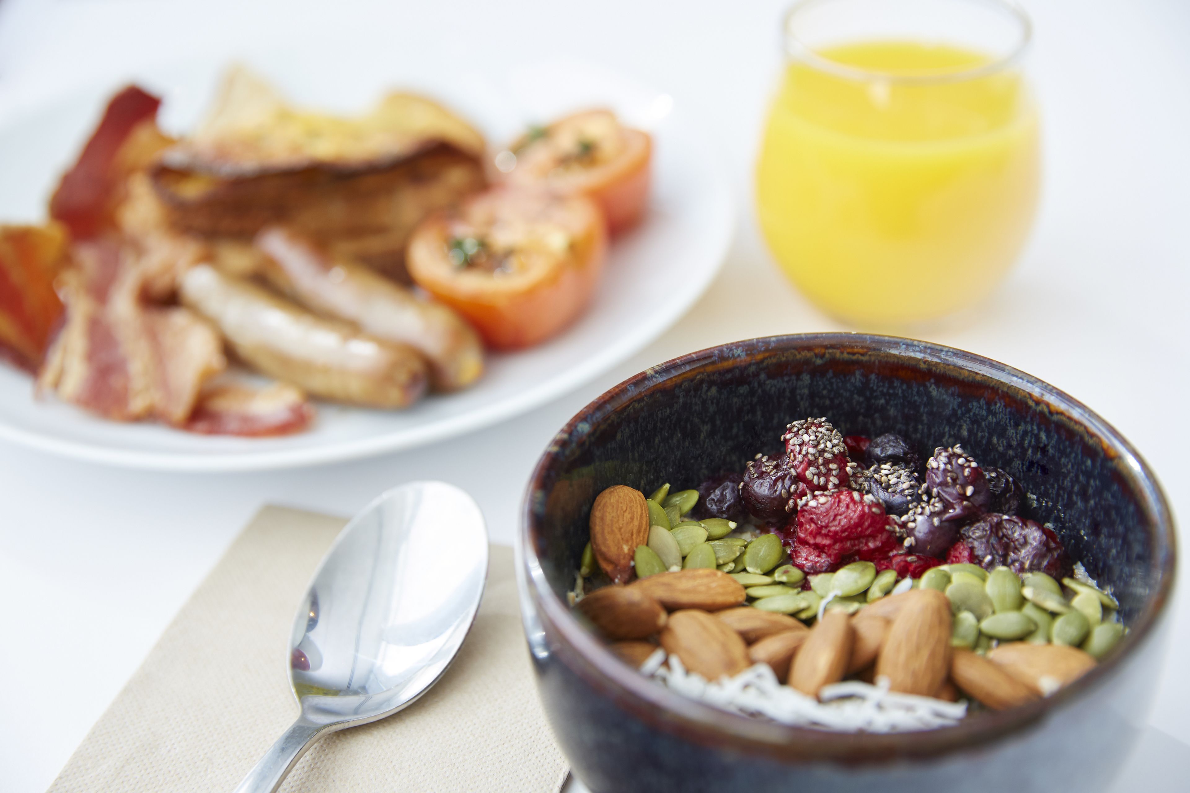 A breakfast setting with a bowl of mixed nuts and seeds, a plate of bacon and eggs, and a glass of orange juice.