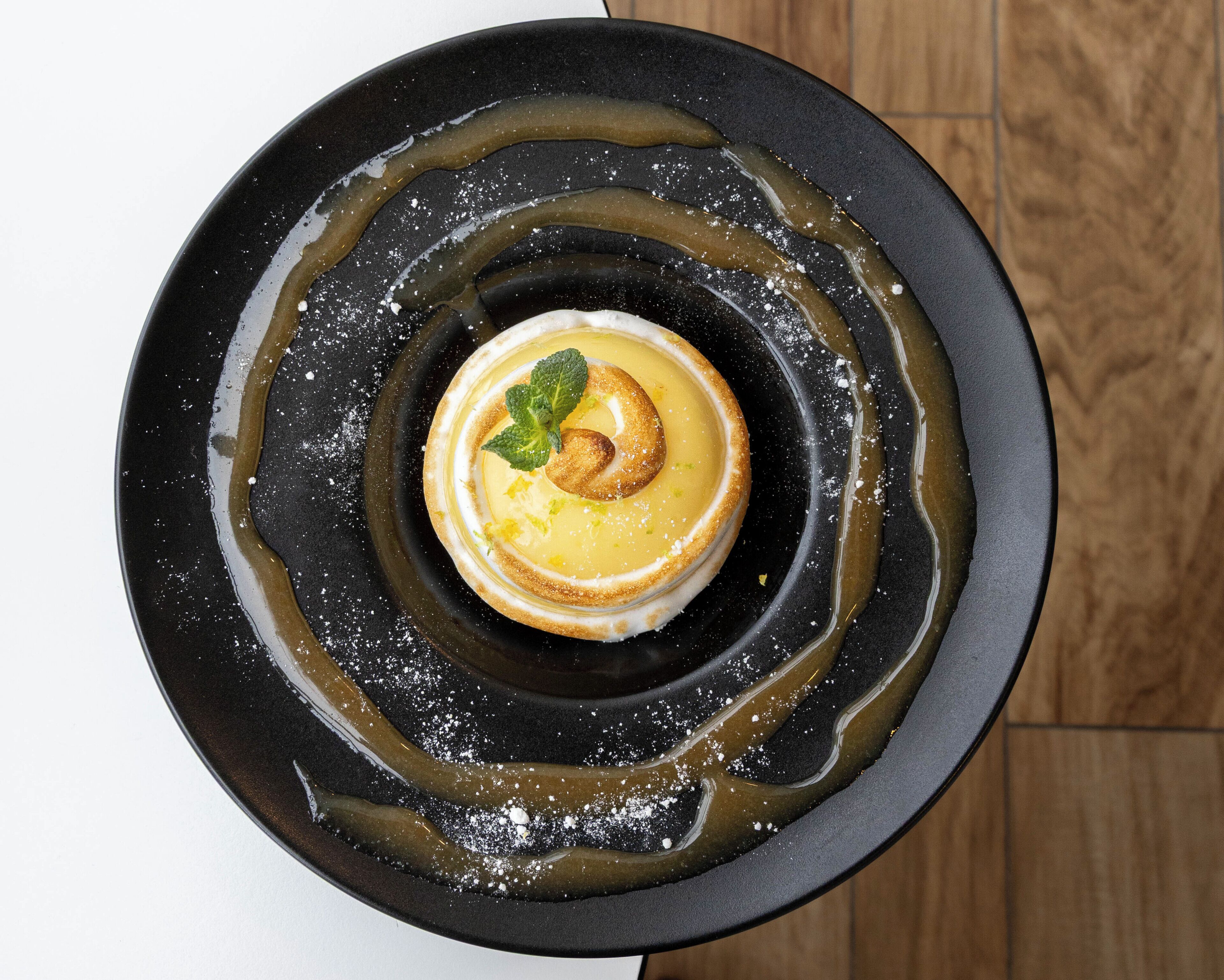 Un postre con una capa caramelizada, una hoja de menta como adorno y una salsa oscura sobre un plato negro.