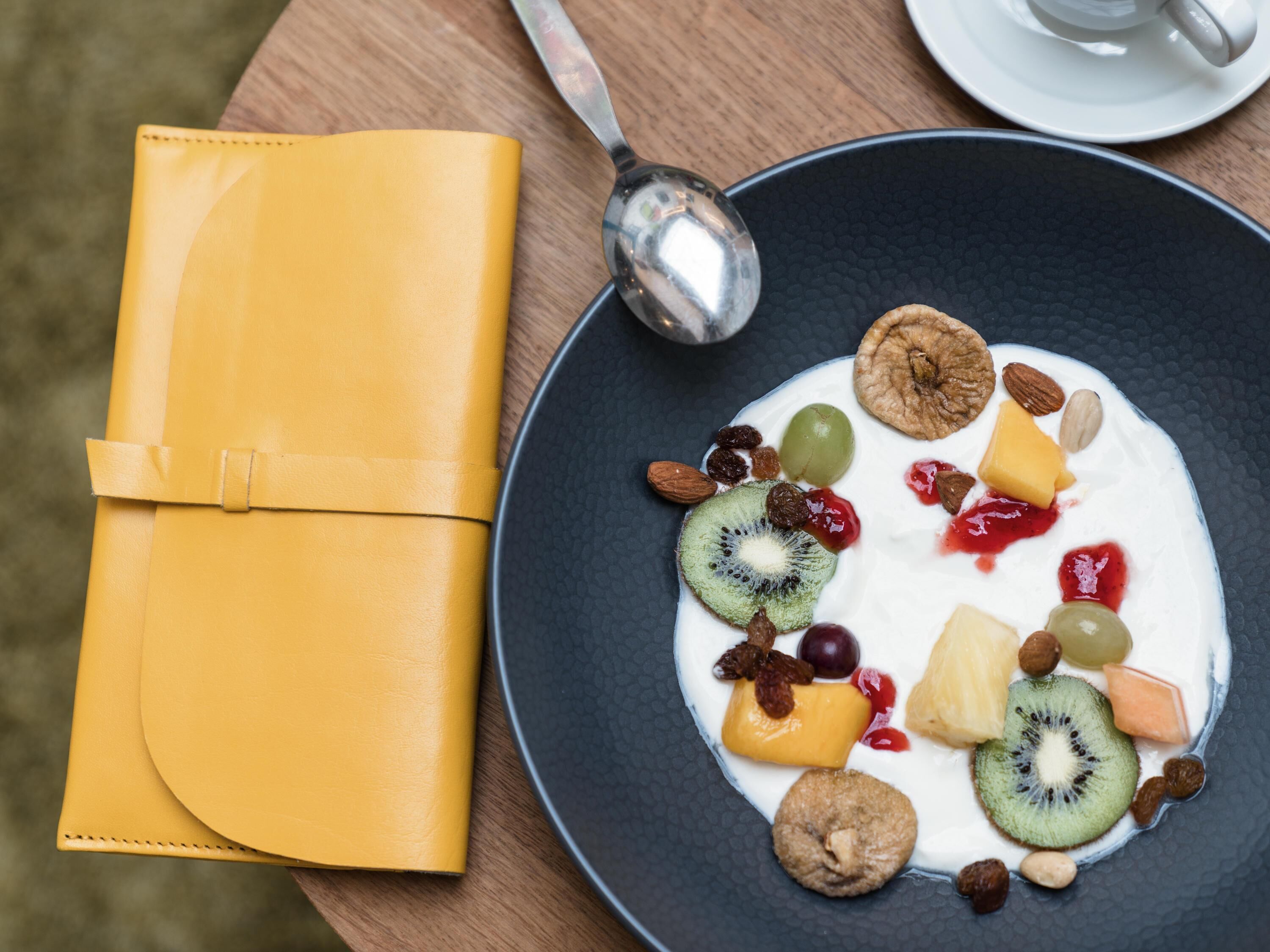A plate of yogurt with fruits and nuts, a spoon, and a yellow napkin on a wooden table.