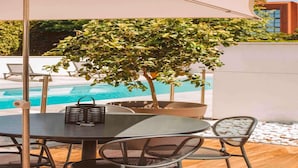 A poolside area with a table and chairs under a white umbrella, surrounded by a tree and a modern building in the background. - Ibis Styles Toulouse Labège (Labege)